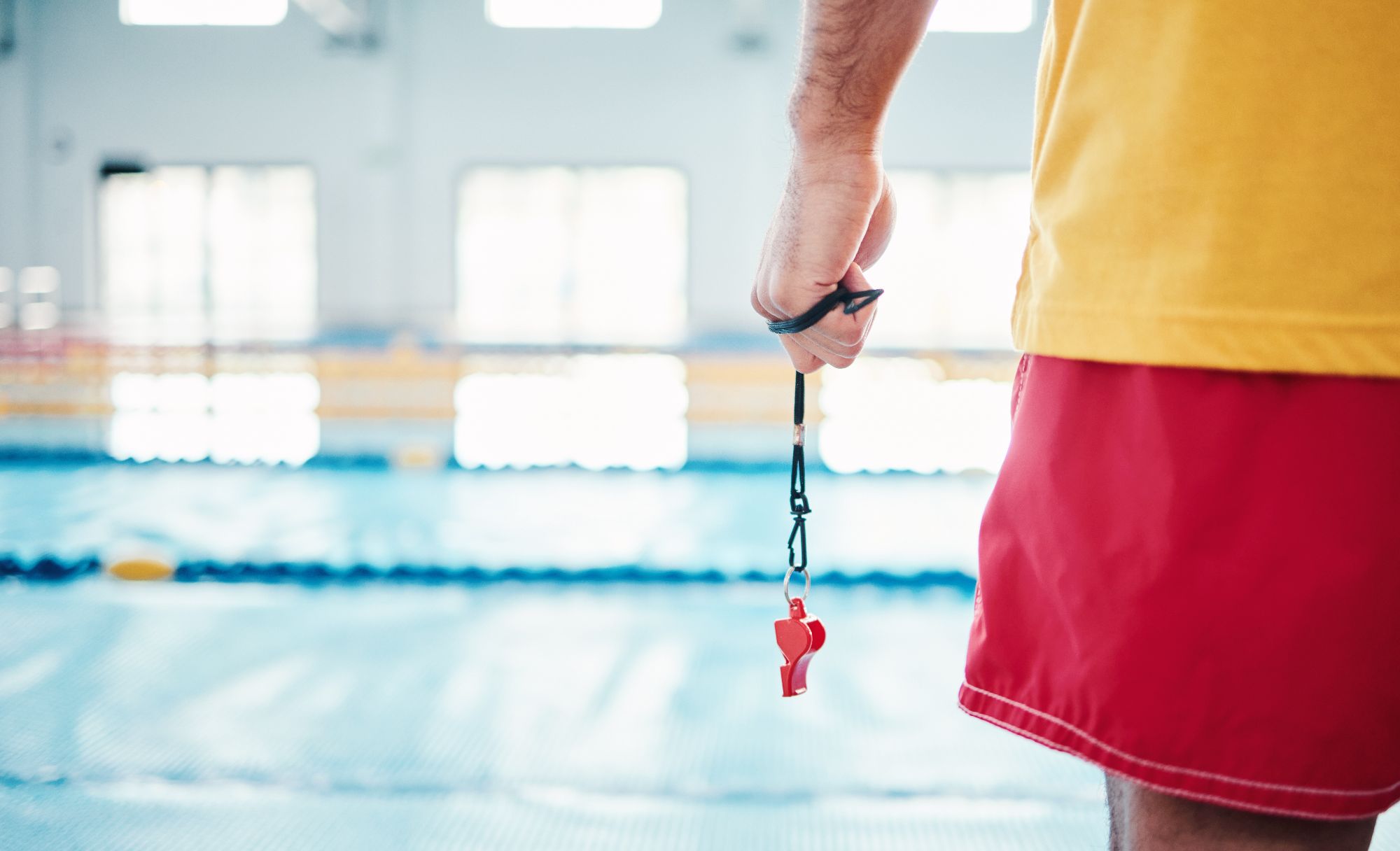 Hands, lifeguard and whistle by swimming pool for water safety, security or ready for rescue indoors. Hand of expert swimmer holding signal tool for warning, safe swim or responsibility for awareness